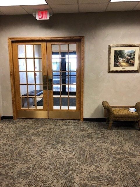 Office hallway with wooden double glass doors, carpeted floor, and a wall bench under framed art.