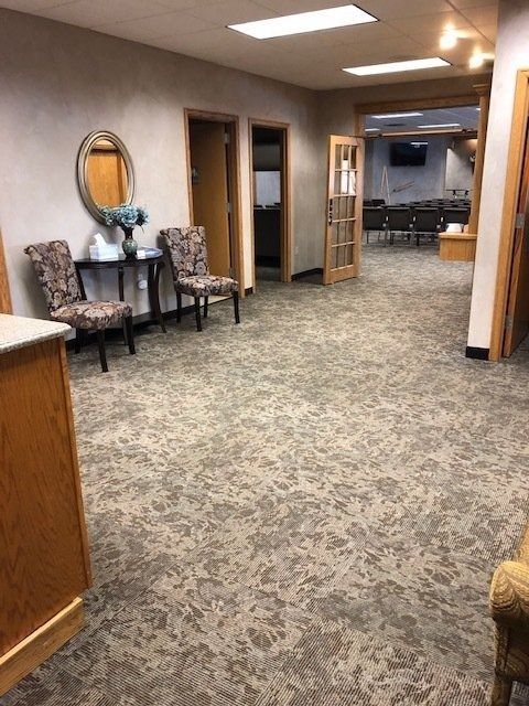 Carpeted hallway with chairs, a wooden cabinet, and open doorways leading to a room in the distance