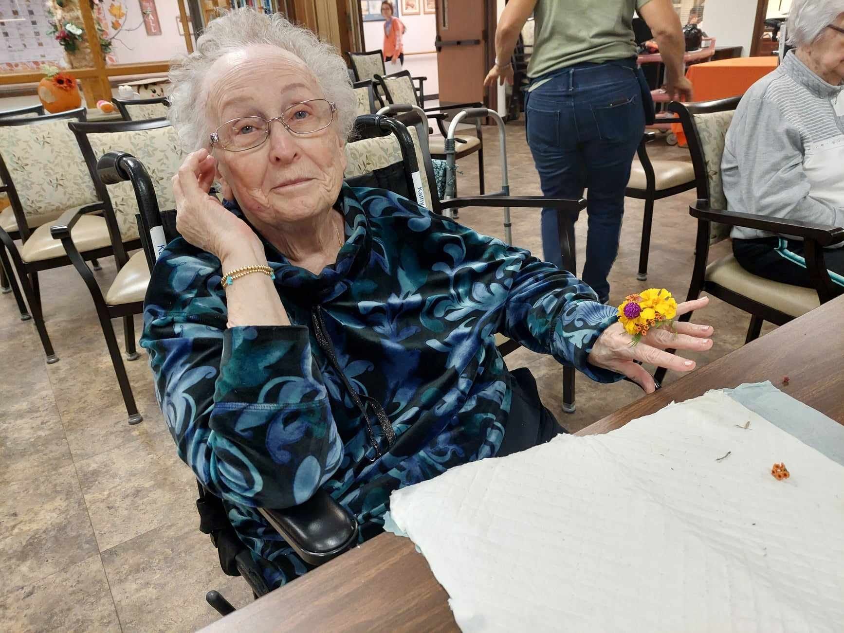 An elderly woman in a wheelchair is sitting at a table holding a flower.