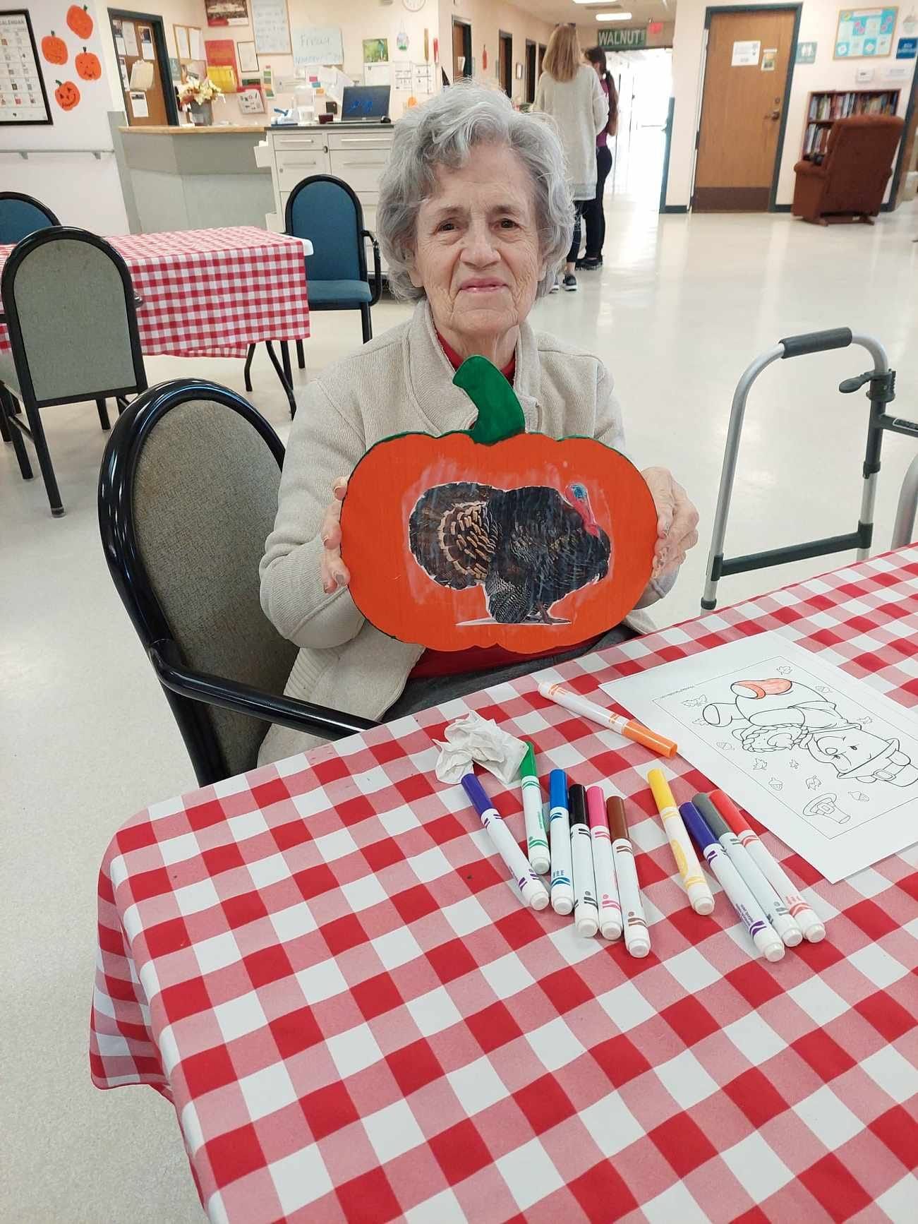 An elderly woman is sitting at a table holding a paper pumpkin