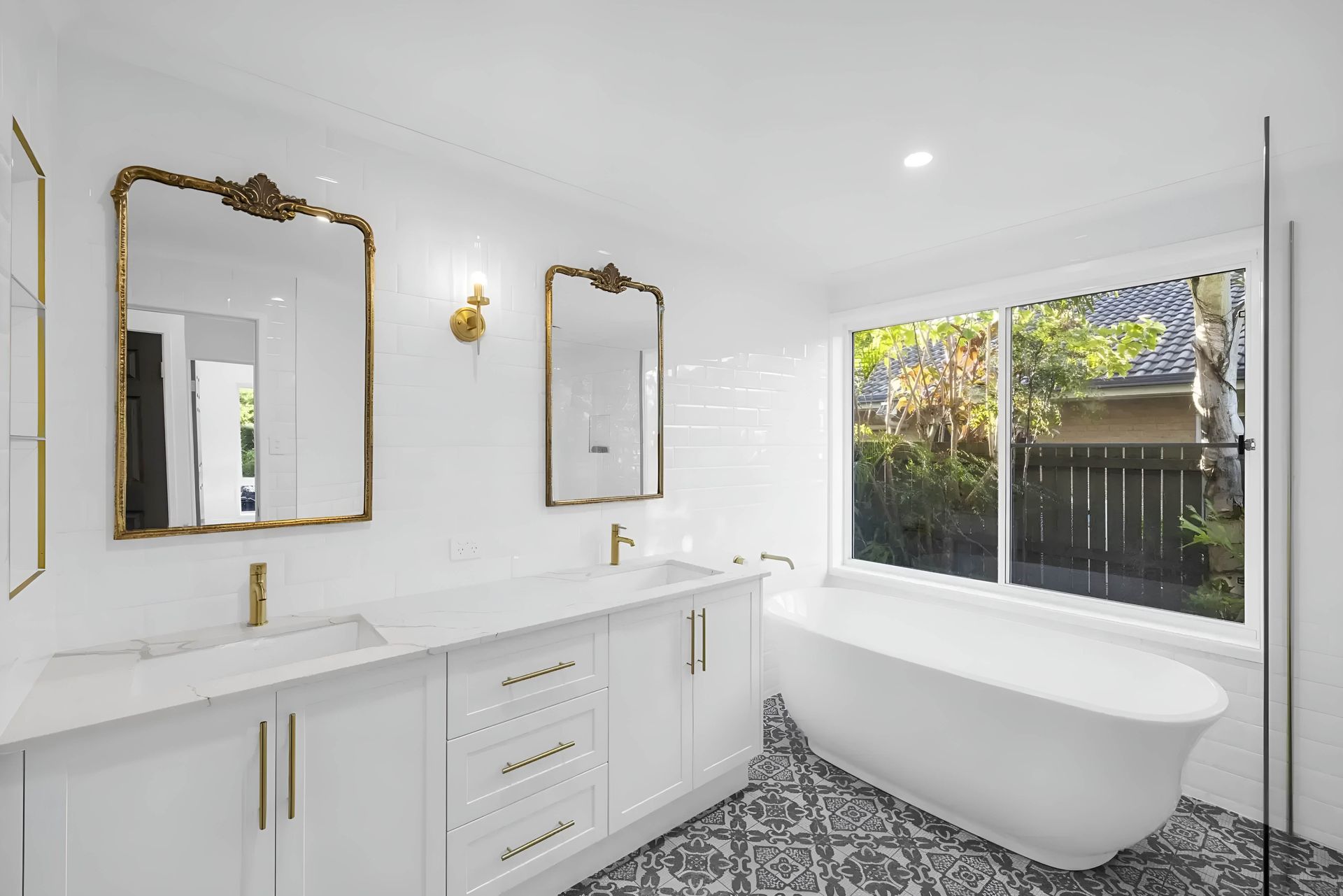 White bathroom with gold accents: double vanity, mirrors, freestanding tub, window with a view, patterned floor