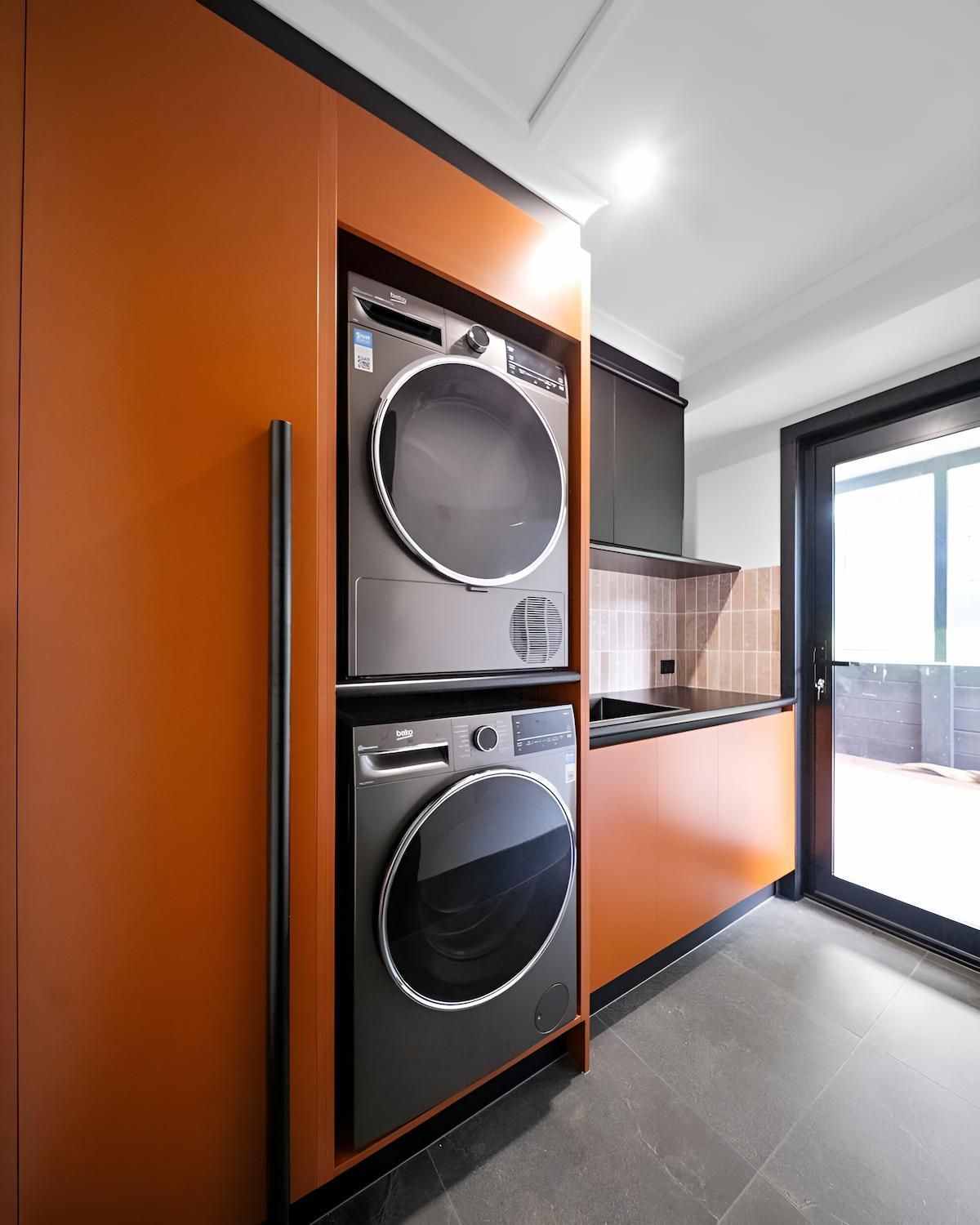Orange cabinetry encloses a stacked washer and dryer. A dark countertop and a glass door are visible