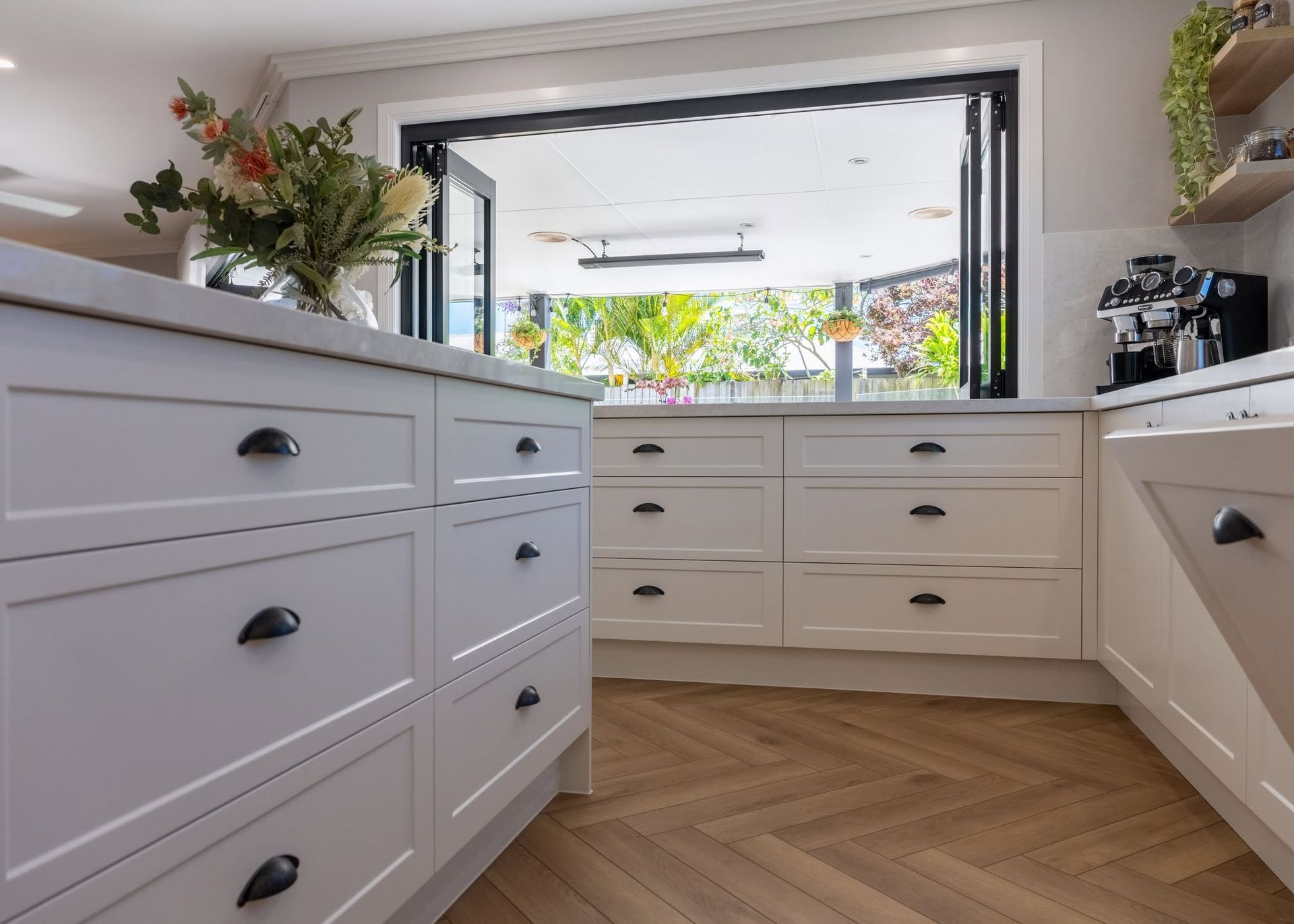 Bright white kitchen with drawers, herringbone wood floor, and an open window with plants outside.