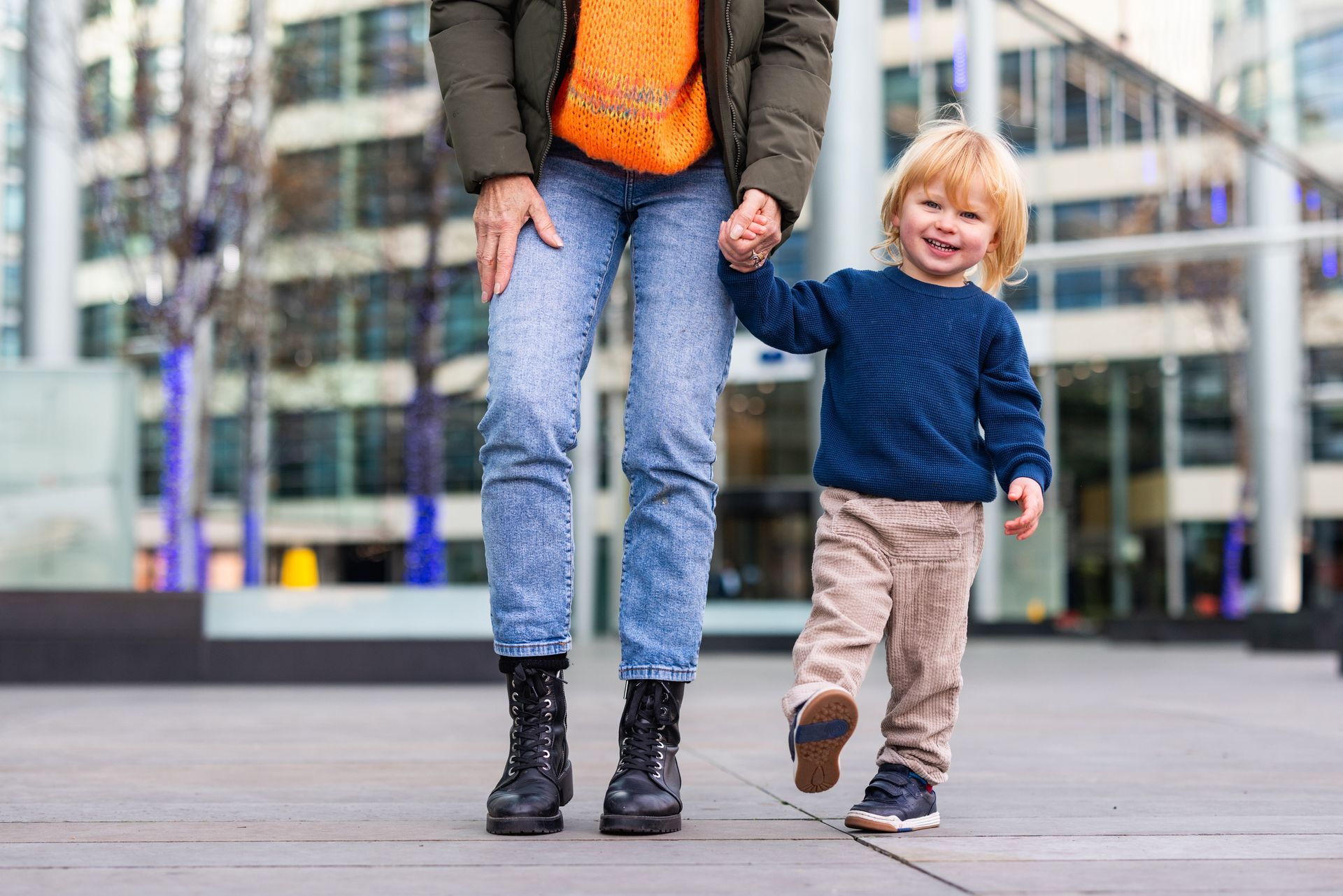 Woman in blue jeans and child in beige pants hold hands, walking outdoors.