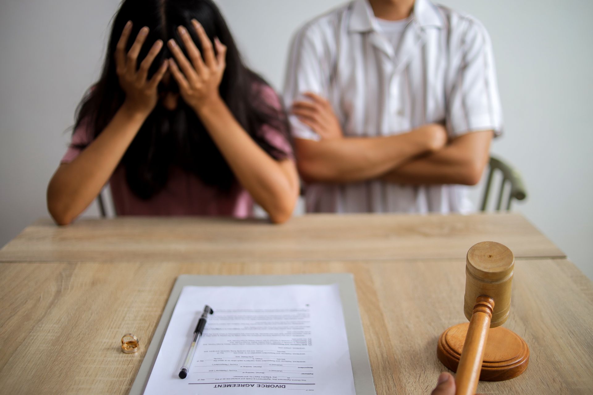 Couple at table, woman with head in hands, legal documents, gavel and ring on table.