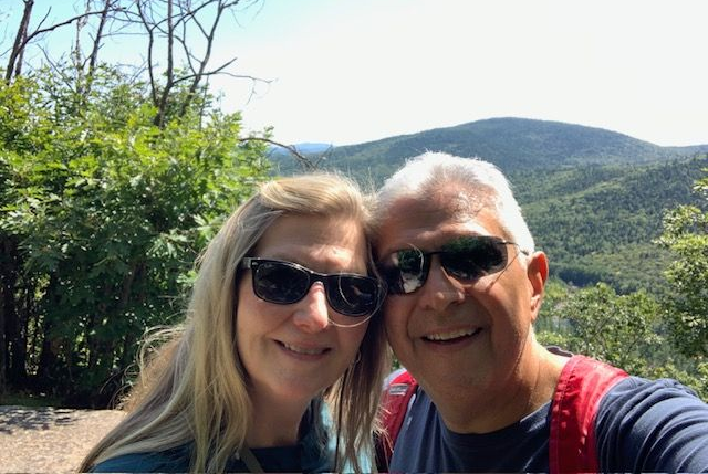 Two smiling people in sunglasses with mountains and greenery behind them