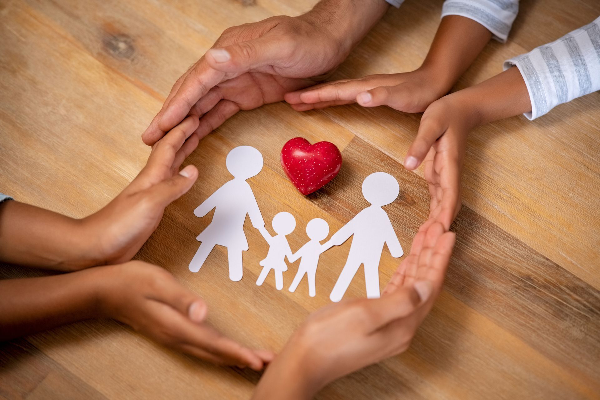 Hands forming a circle around paper family figures and a red heart on a wooden table