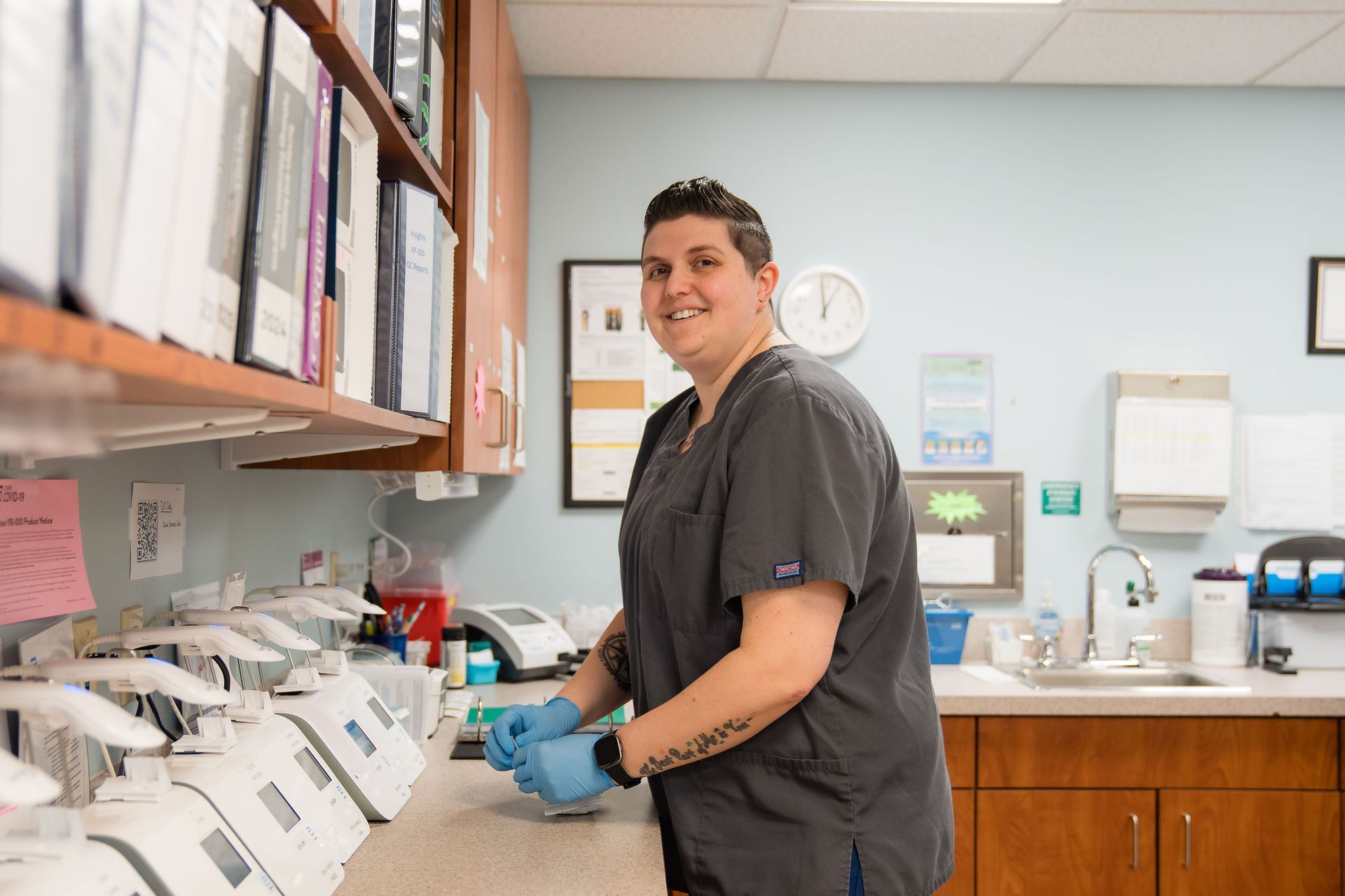 A lab tech is standing in a lab wearing scrubs and gloves.