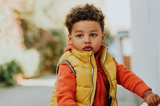 Young child in a yellow vest and orange sleeves standing outdoors against a blurred background