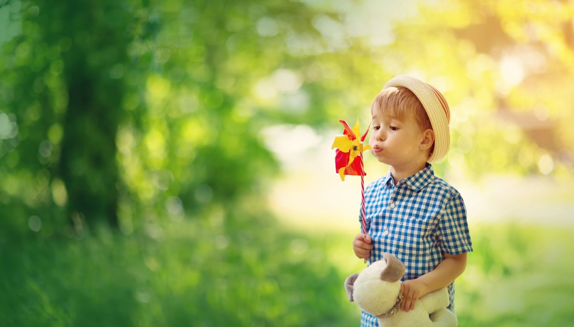 A little boy is blowing a windmill in the park while holding a teddy bear.