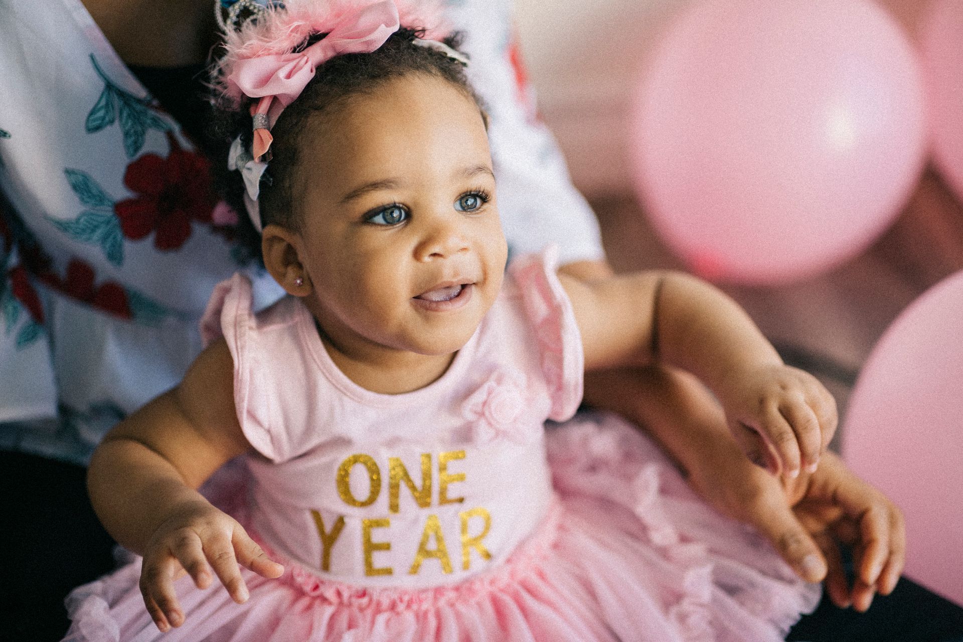 A baby girl wearing a pink dress and headband is sitting next to a woman.
