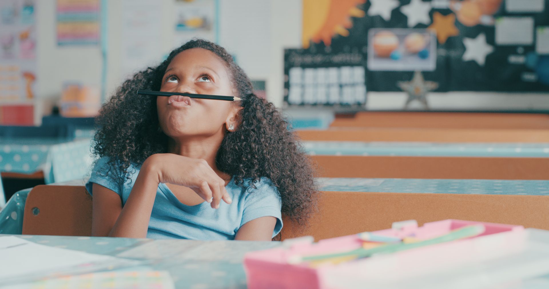 A young girl is sitting at a desk in a classroom with a pencil in her mouth.