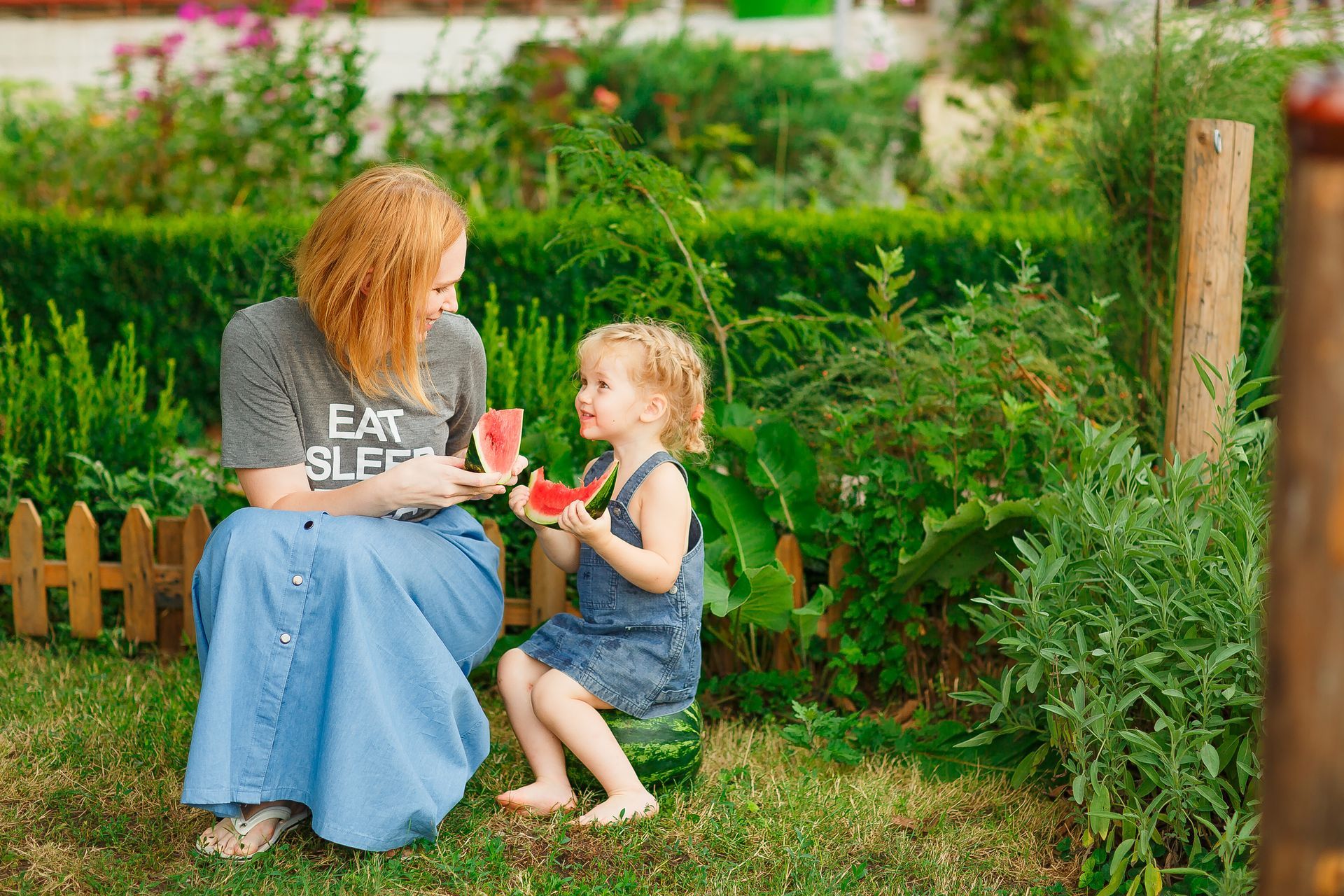 A woman and a little girl are eating watermelon in a garden.