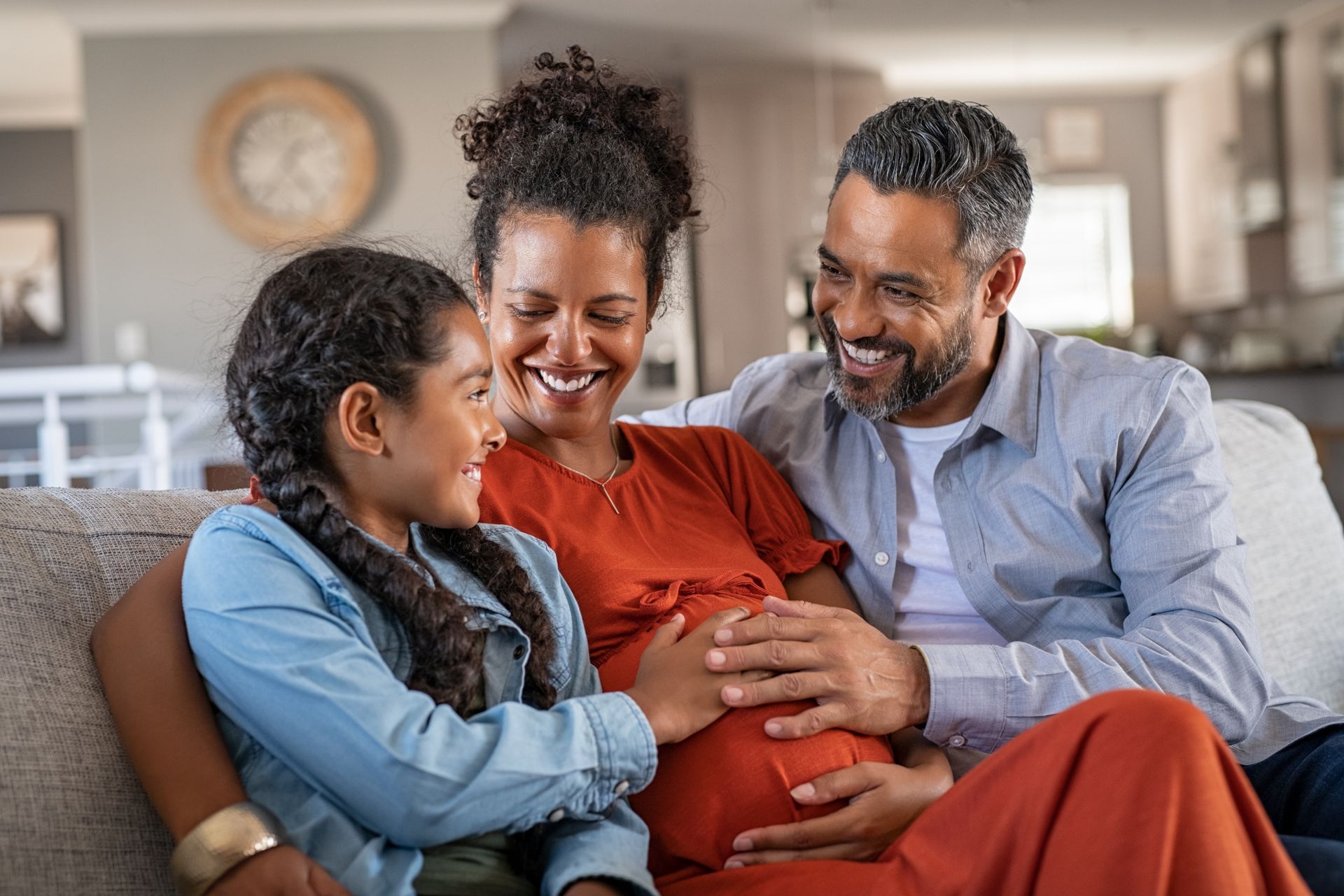A pregnant woman is sitting on a couch with her husband and daughter.