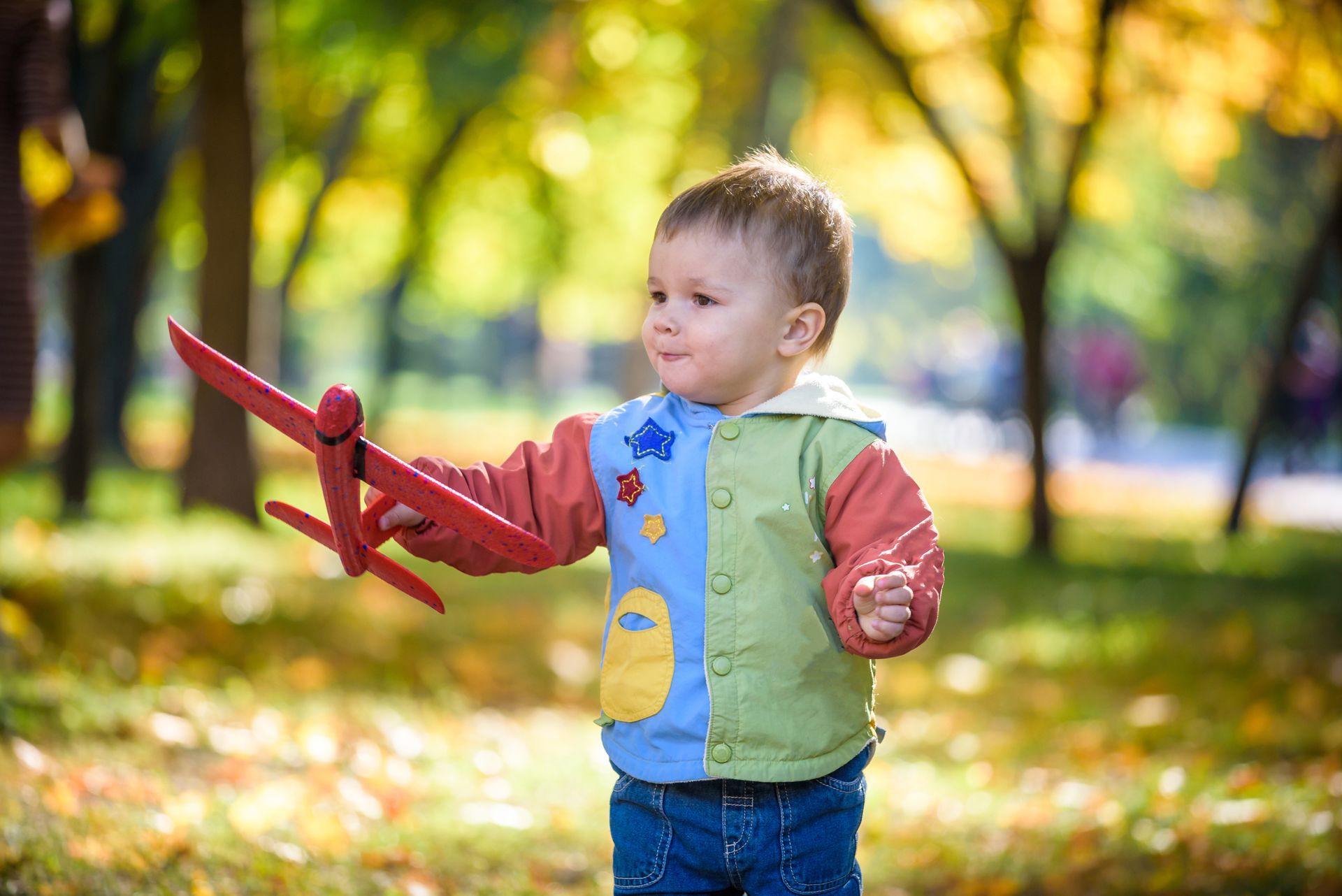 Young child holds a red toy airplane in a park with autumn foliage.