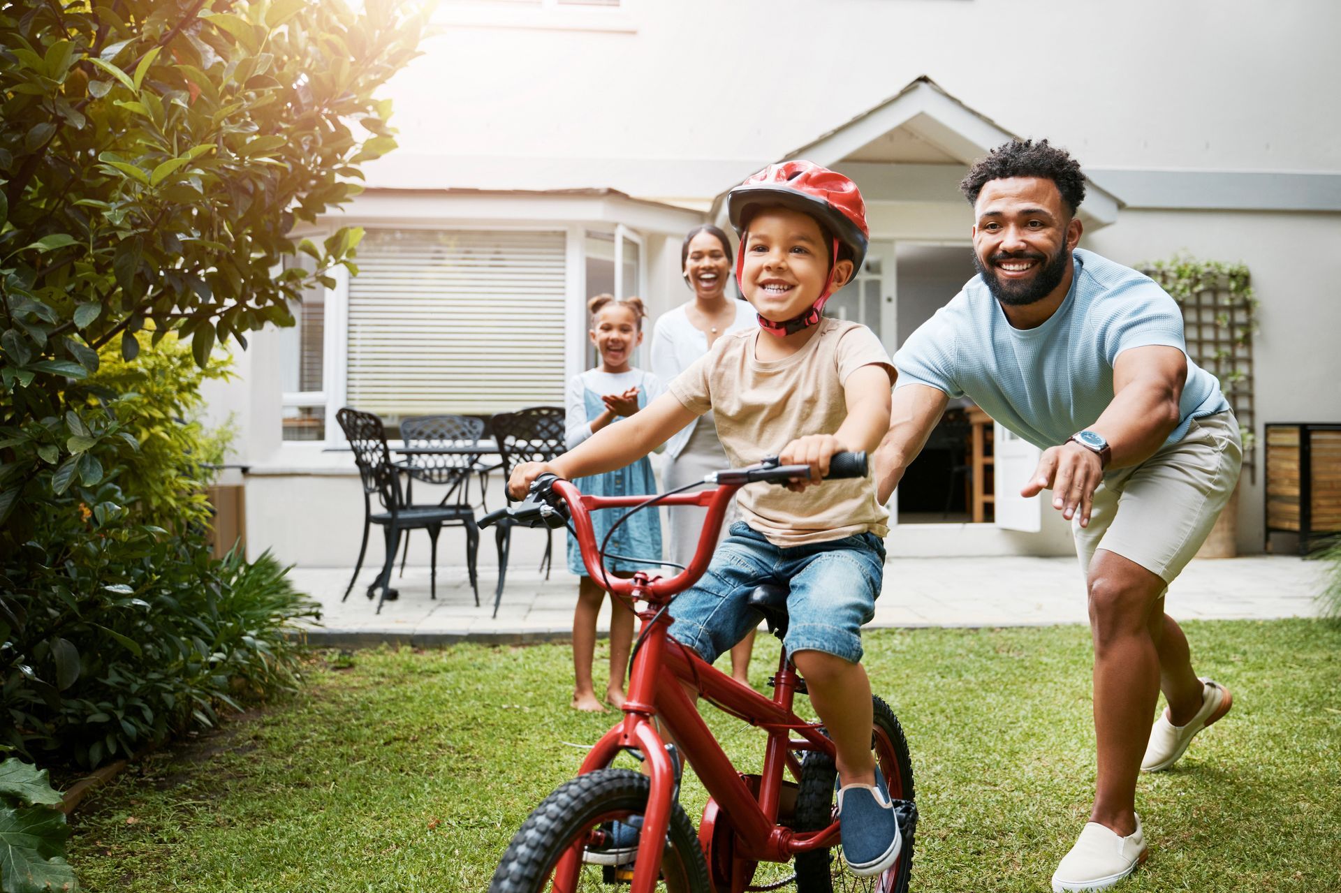 A family watching as a young boy learns to ride a red bicycle