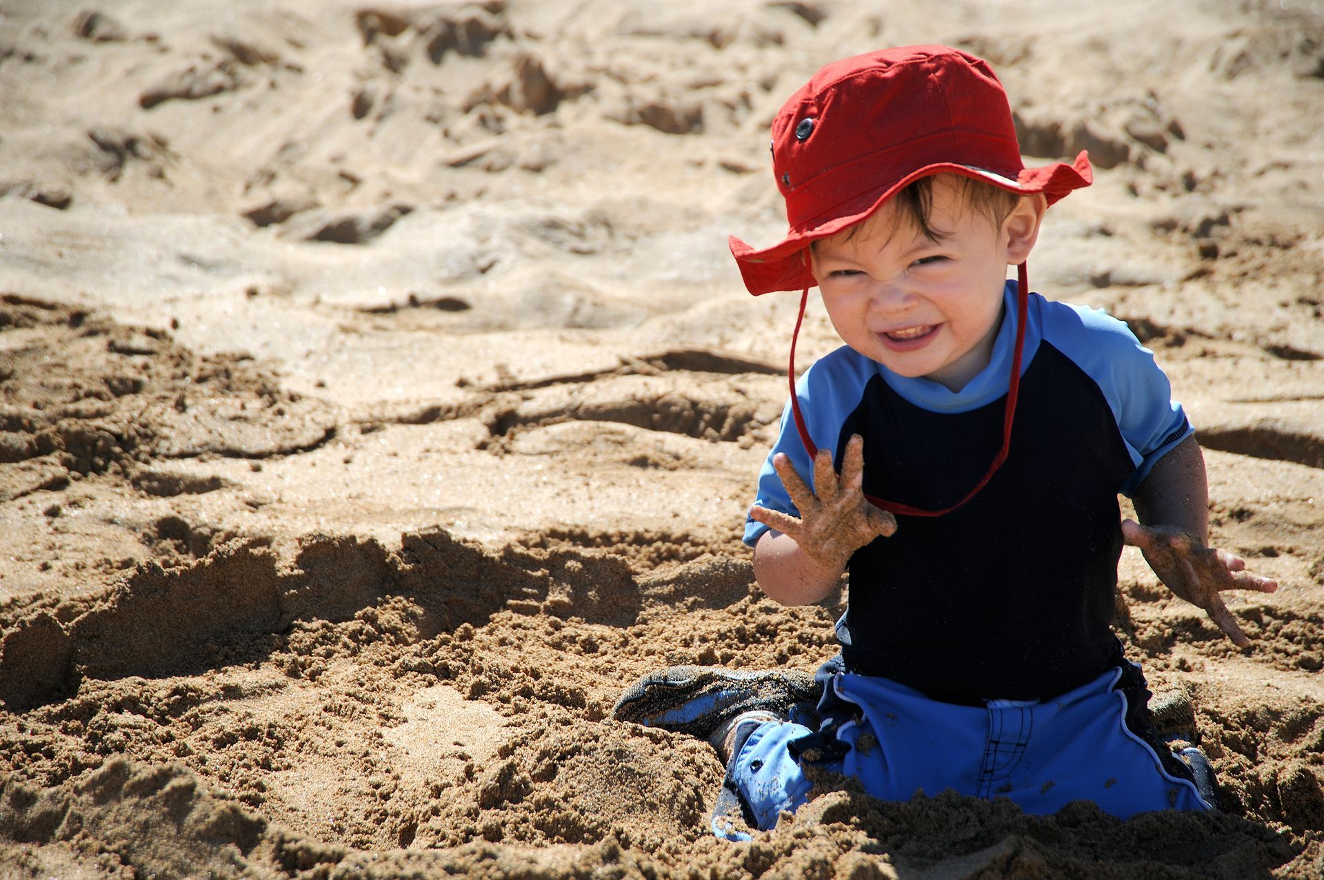 A little boy wearing a red hat is playing in the sand