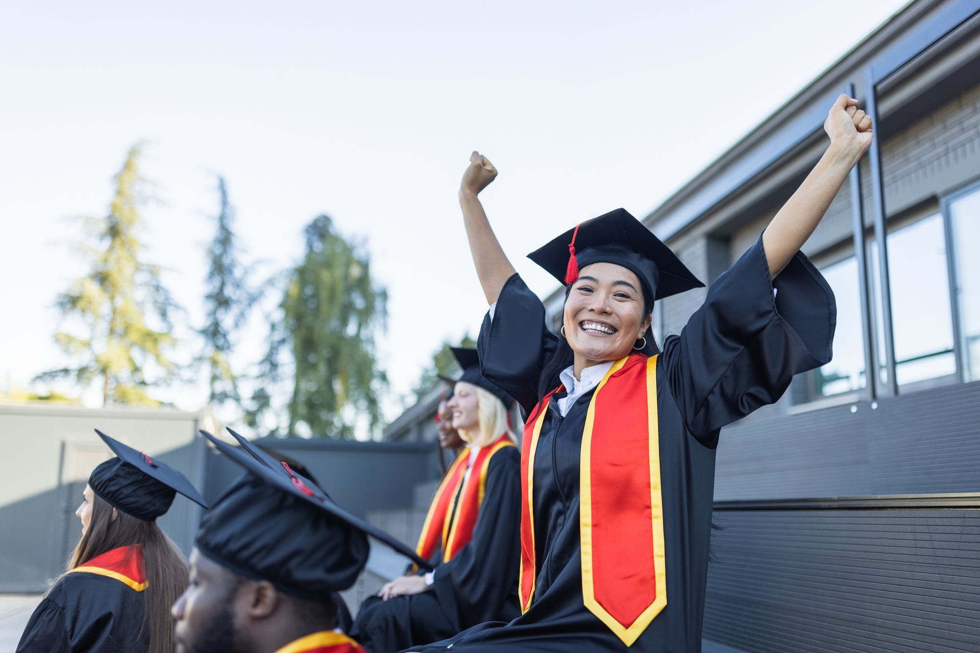 A group of graduates are sitting on the steps of a building with their arms in the air.