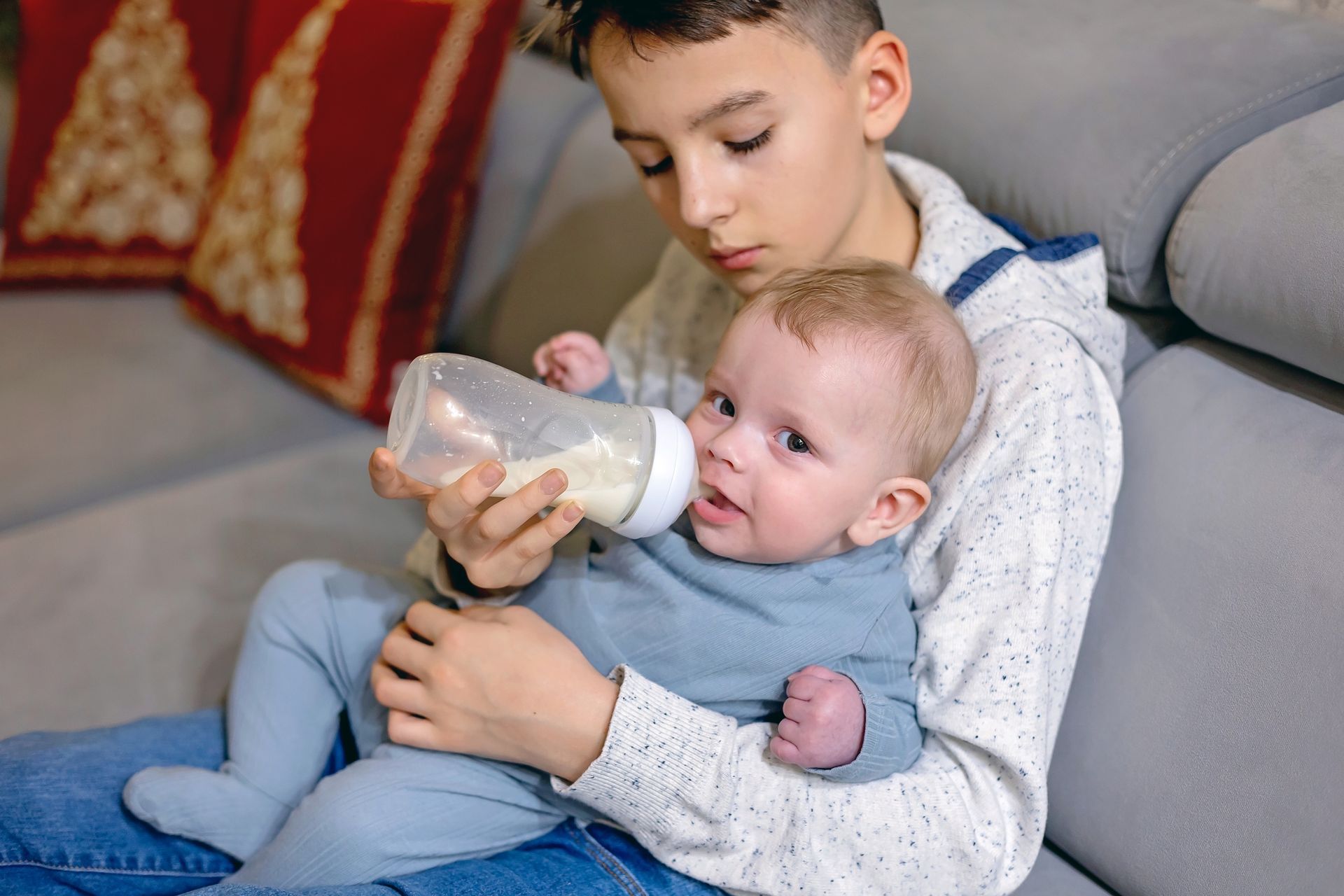 A young boy is feeding a baby from a bottle while sitting on a couch.
