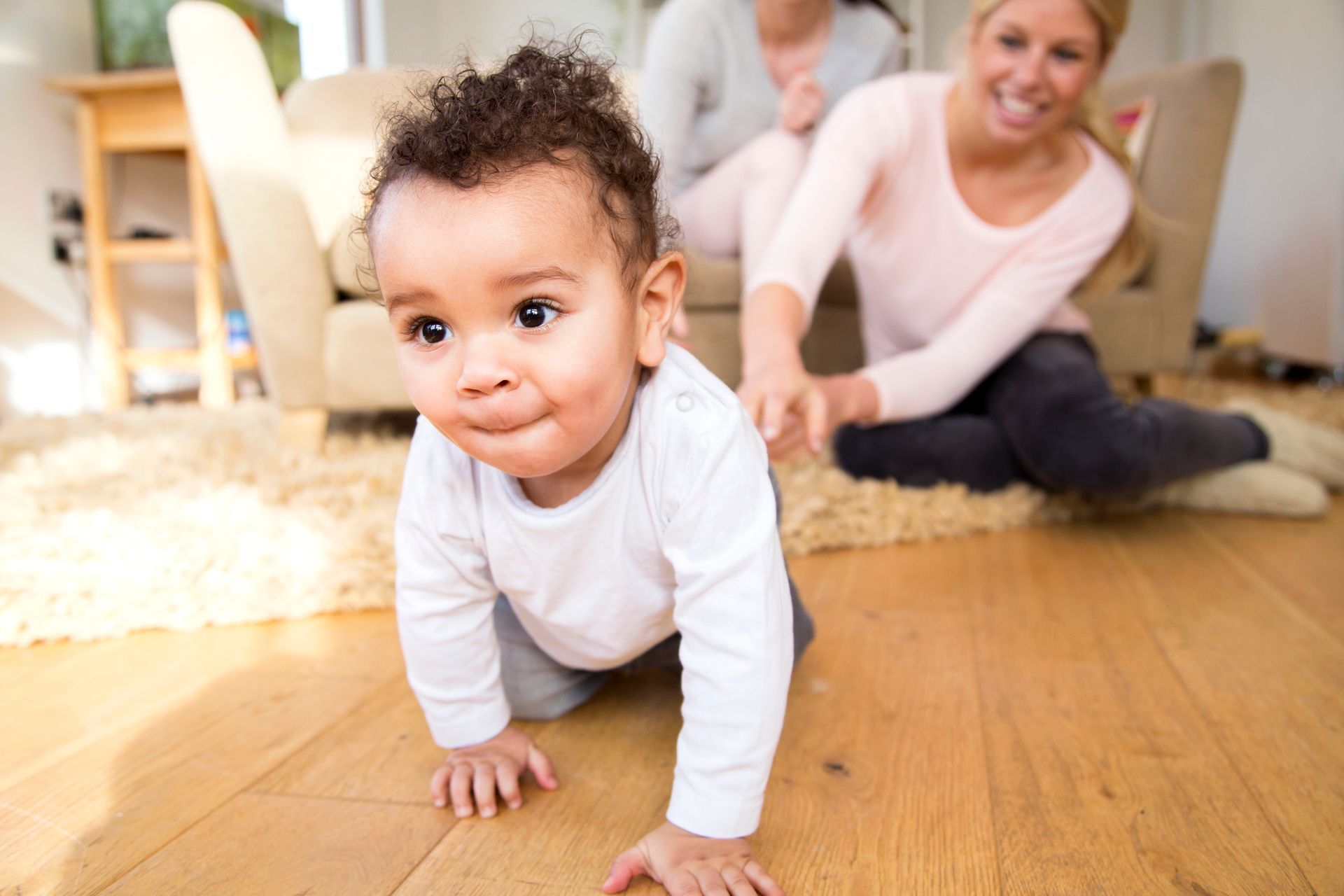 A baby is crawling on the floor in a living room while a woman sits on the floor.