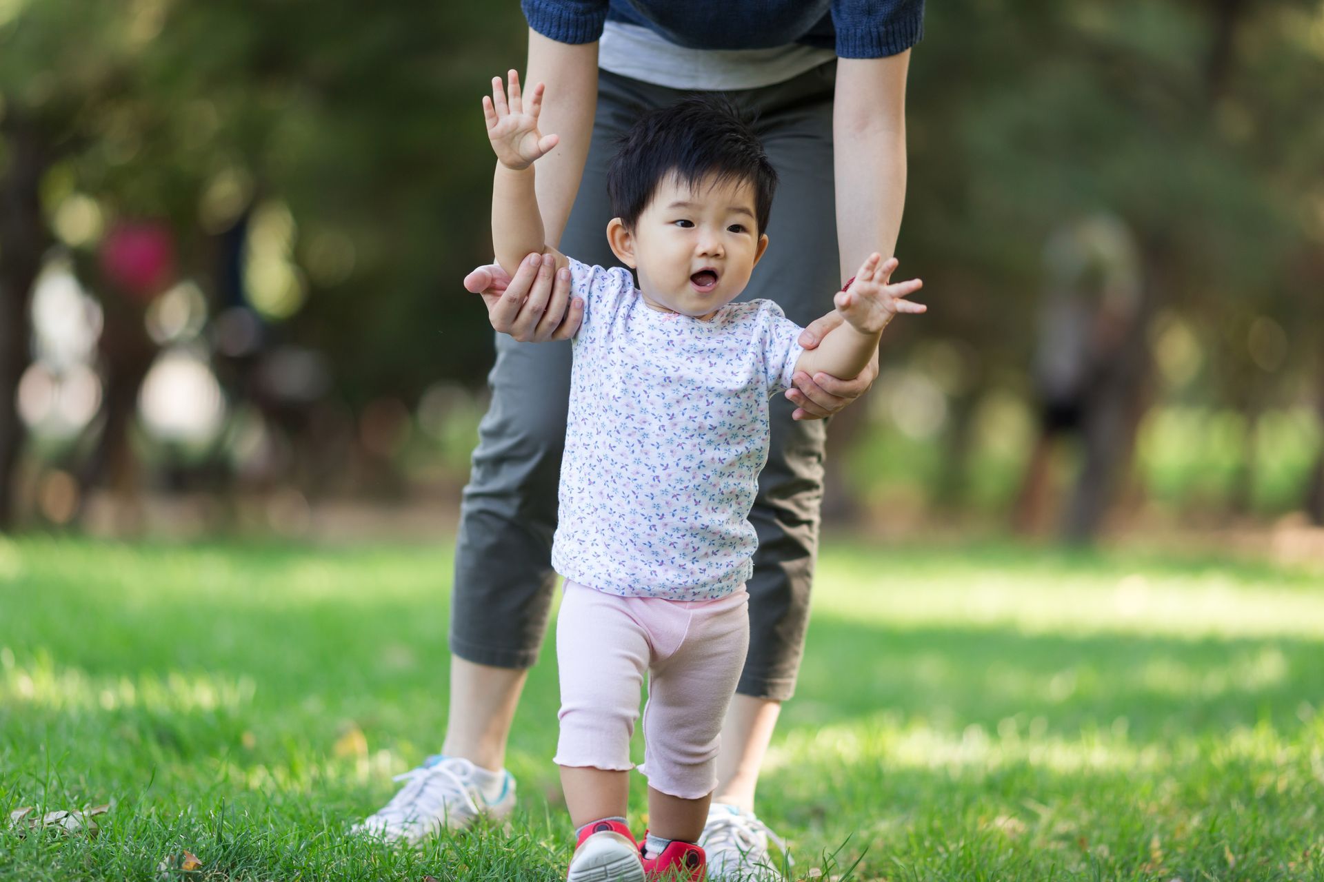 A woman is helping a baby girl walk in a park.