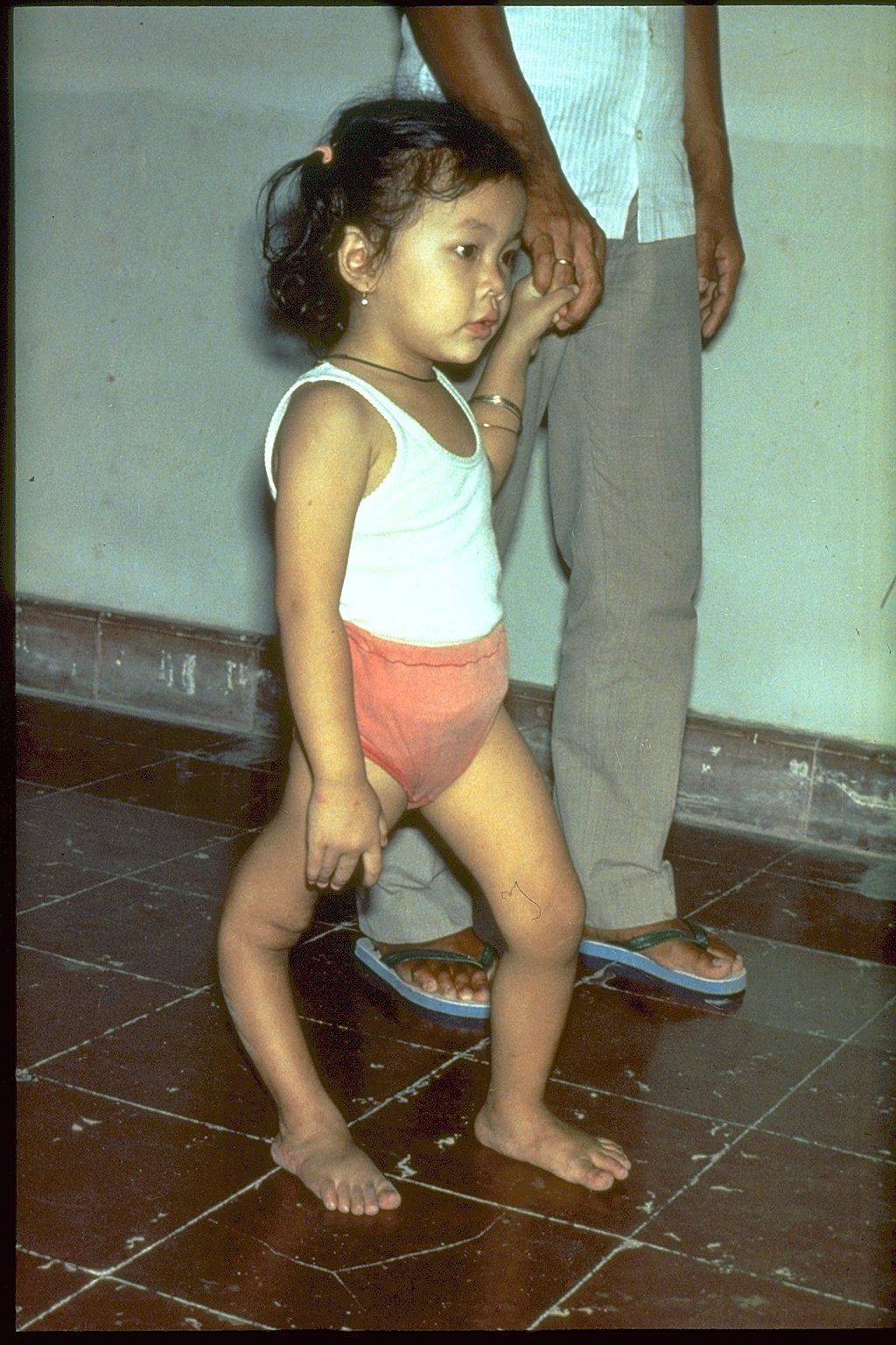 Small child in a white tank top and orange shorts walking indoors beside an adult.