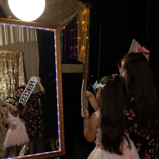 People taking selfies in a photo booth with a mirror, string lights, and gold sequin curtains
