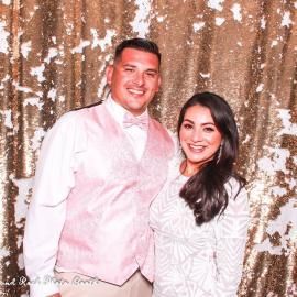 A smiling couple poses in white attire before a gold sequin backdrop.