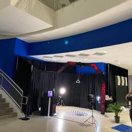 Indoor event space with a black stage backdrop, blue walls, lighting equipment, and stairs on the left