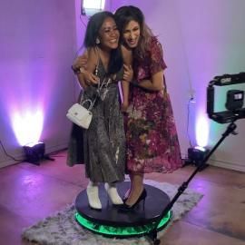 Two women posing on a lit rotating platform in a photo booth, smiling and hugging.
