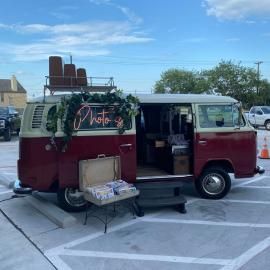 Vintage maroon-and-white VW bus food truck with open side door and flower decorations in a parking lot