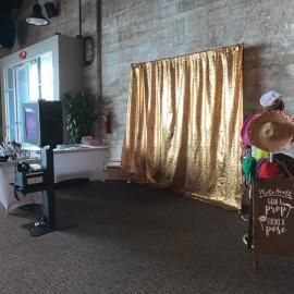 Indoor event space with gold curtain backdrop, TV on a cart, and a decorative sign with flowers.