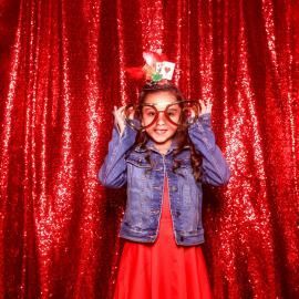Child in denim jacket and red dress holding oversized glasses in front of a red sequin backdrop