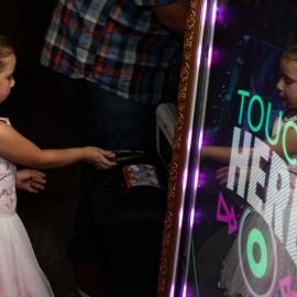 Child reaching toward a glowing touch screen with neon “TOUCH HERE” text.