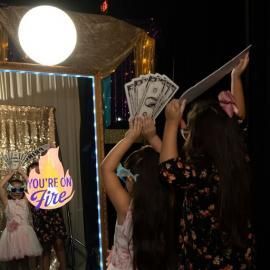 People celebrating at a photo booth, one holding cash and another lifting a prop against a glittery backdrop