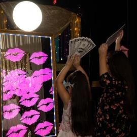 Two people posing by a pink lip backdrop, one holding cash and the other a phone, at a party