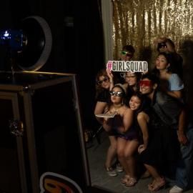 Group posing at a party beside a #GIRLSQUAD sign and gold backdrop, with a camera light on the left.