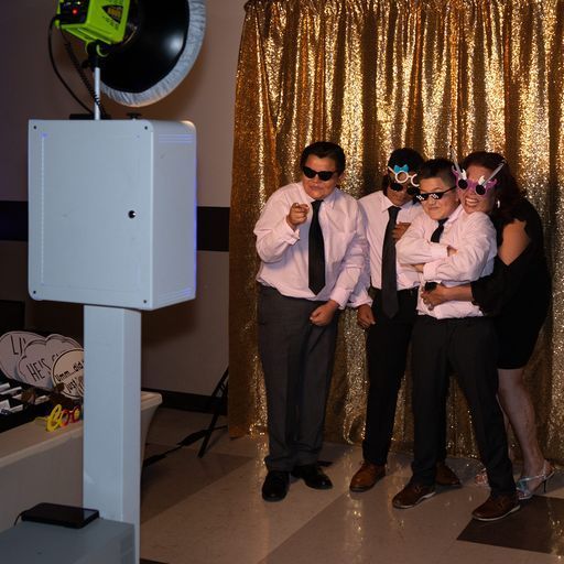 Four people in sunglasses posing at a photo booth with a gold curtain backdrop