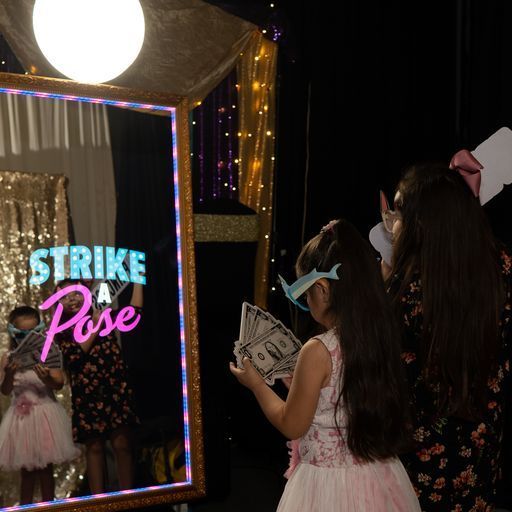Two girls posing at a neon-lit “Strike a Pose” photo booth holding props