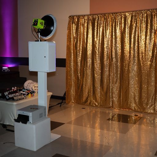 Photo booth kiosk with a ring light and green camera in a room beside a gold curtain backdrop