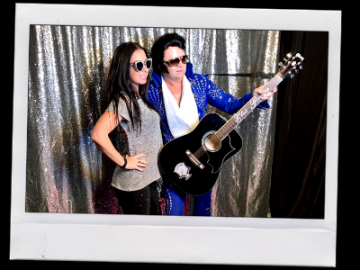 Two people posing in sunglasses, one in a blue jacket holding a black guitar, in front of a silver curtain