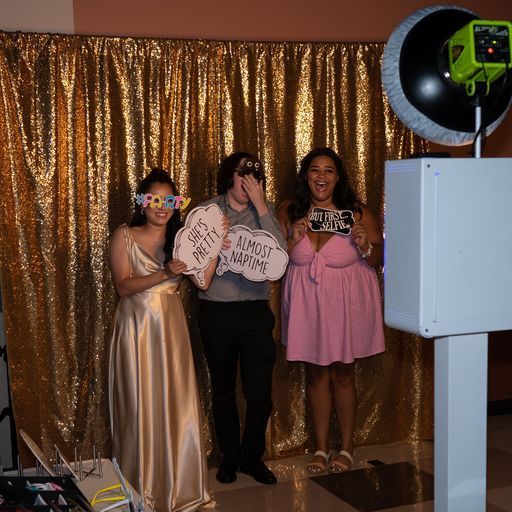 Three people posing in front of a gold sequin backdrop at a photo booth, holding signs and wearing party outfits.