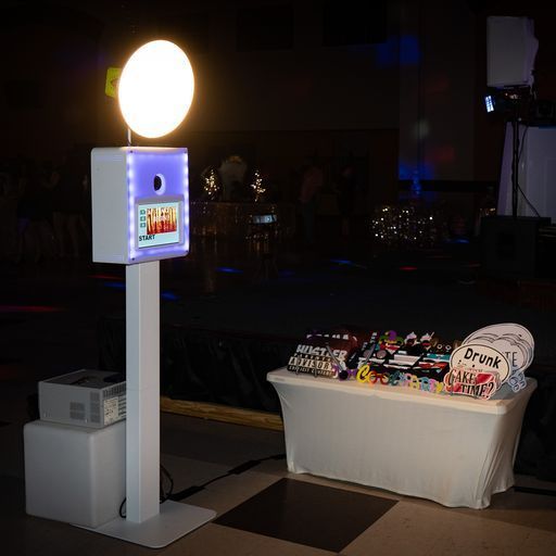 Photo booth setup with glowing circular light, white kiosk, and prize table in a dark event space
