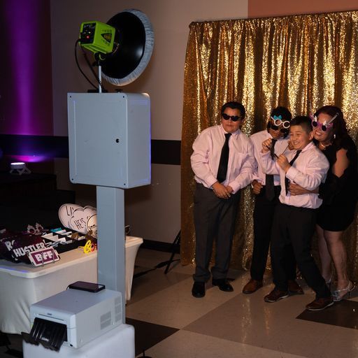 Four people pose beside a photo booth with a gold curtain backdrop at a party.