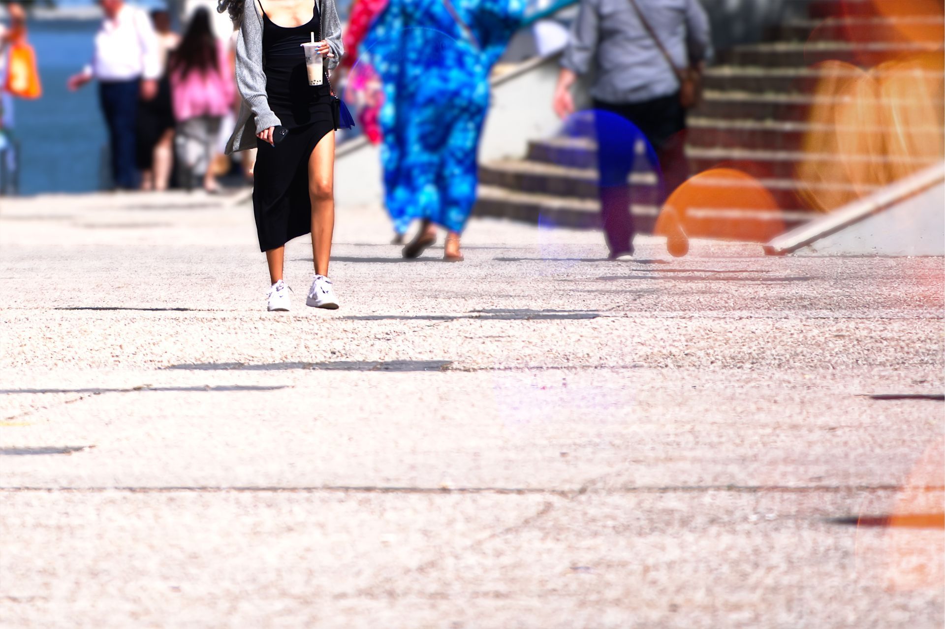 People walking on a concrete path near water; sunny day. Woman in black dress, others blurred in the background.