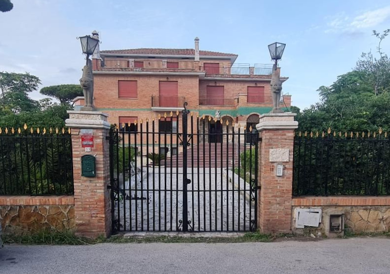 Ingresso recintato di una villa a due piani in mattoni con facciata rossa e recinzione in ferro nero sotto un cielo azzurro