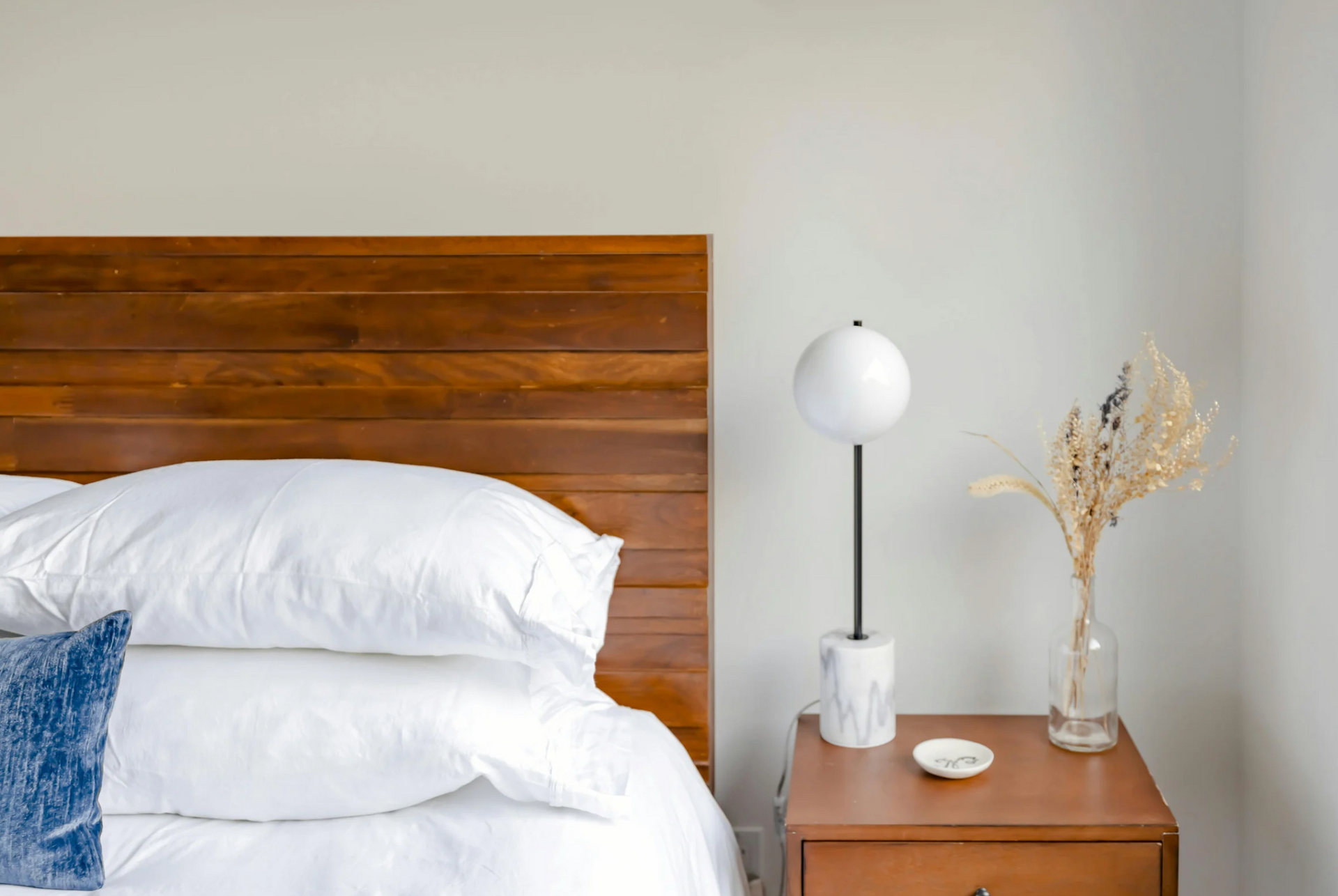 A bedroom with a bed , nightstand , lamp and vase of dried flowers.