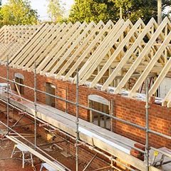 Bâtiment en briques en construction avec fermes de toit en bois apparentes et échafaudages.