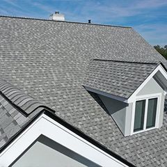Toit en bardeaux gris d'une maison avec une lucarne. Ciel bleu visible en arrière-plan.