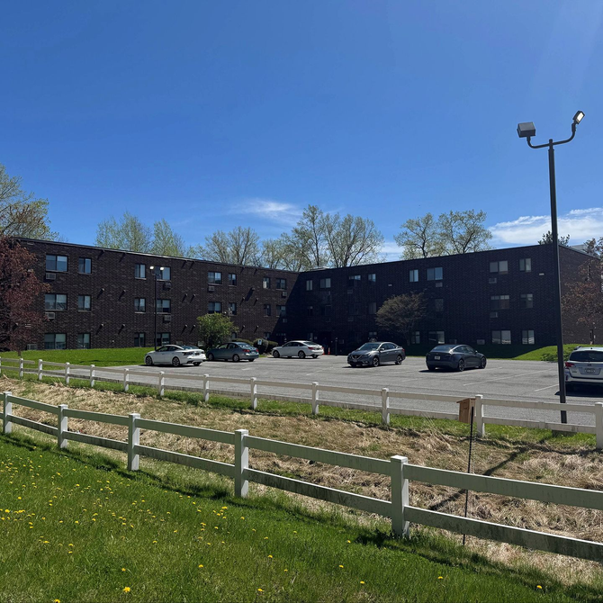 Brick apartment building beside a parking lot and grassy field under a clear blue sky